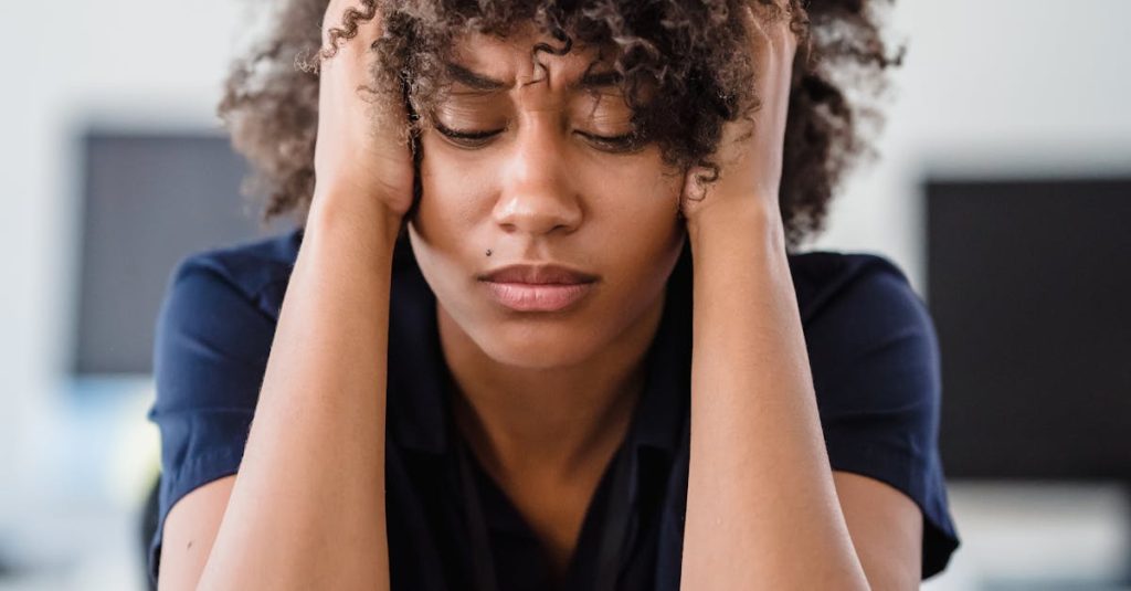 Woman in office setting expresses stress, seated at desk with hands in hair. Perfect for workplace stress concept.