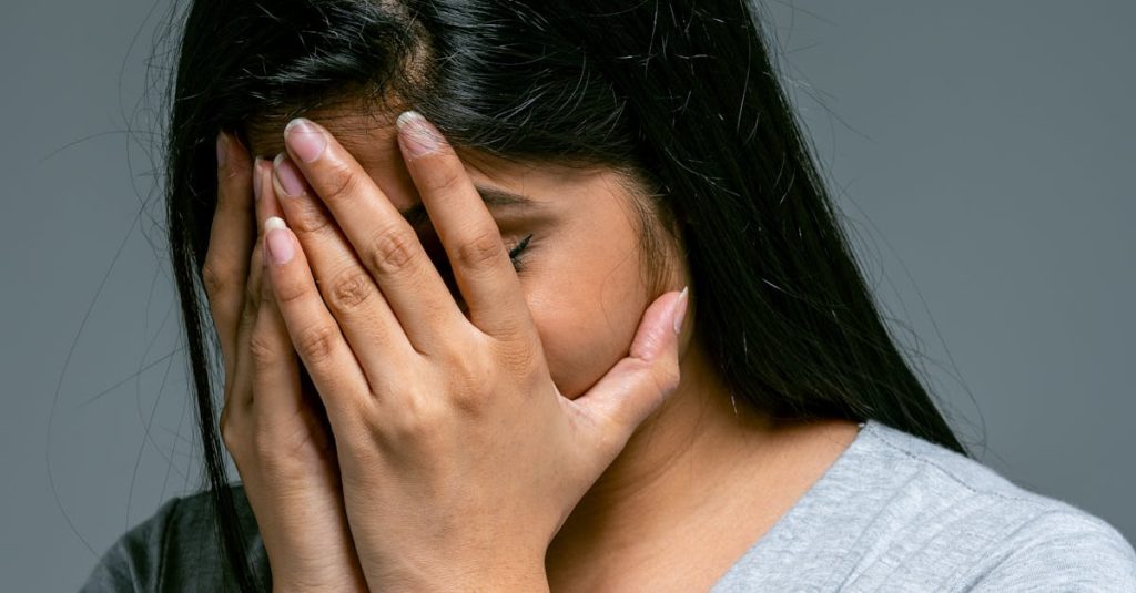 A woman in a gray shirt hides her face in hands expressing stress and emotion.