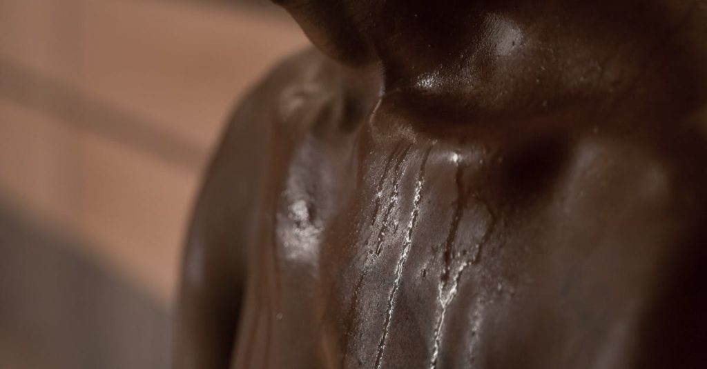 Close-up of a shirtless man in dim light, highlighting his skin with shadows and sweat.