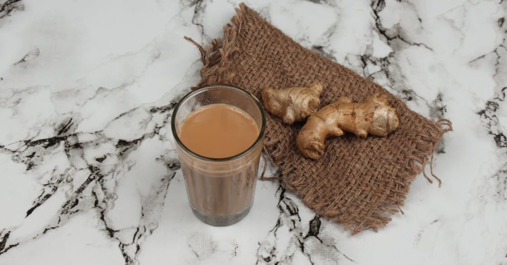 A glass of traditional Indian masala chai served with fresh ginger on a marble countertop.