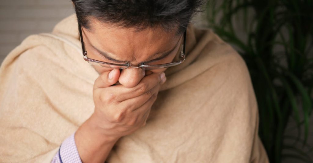 Adult wearing glasses coughs into hand indoors, indicating illness or flu symptoms.