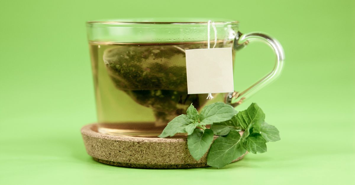 Green tea bag in glass cup with hot water on cup holder with mint leaves placed on green background