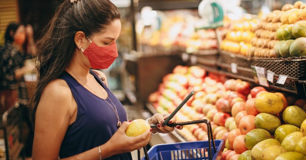 A woman wearing a mask selects fresh fruits in a supermarket produce aisle.